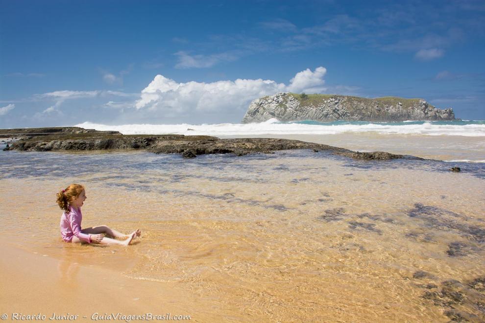 Imagem de uma menina sentada na beira do mar rindo para o mar maravilhoso.
